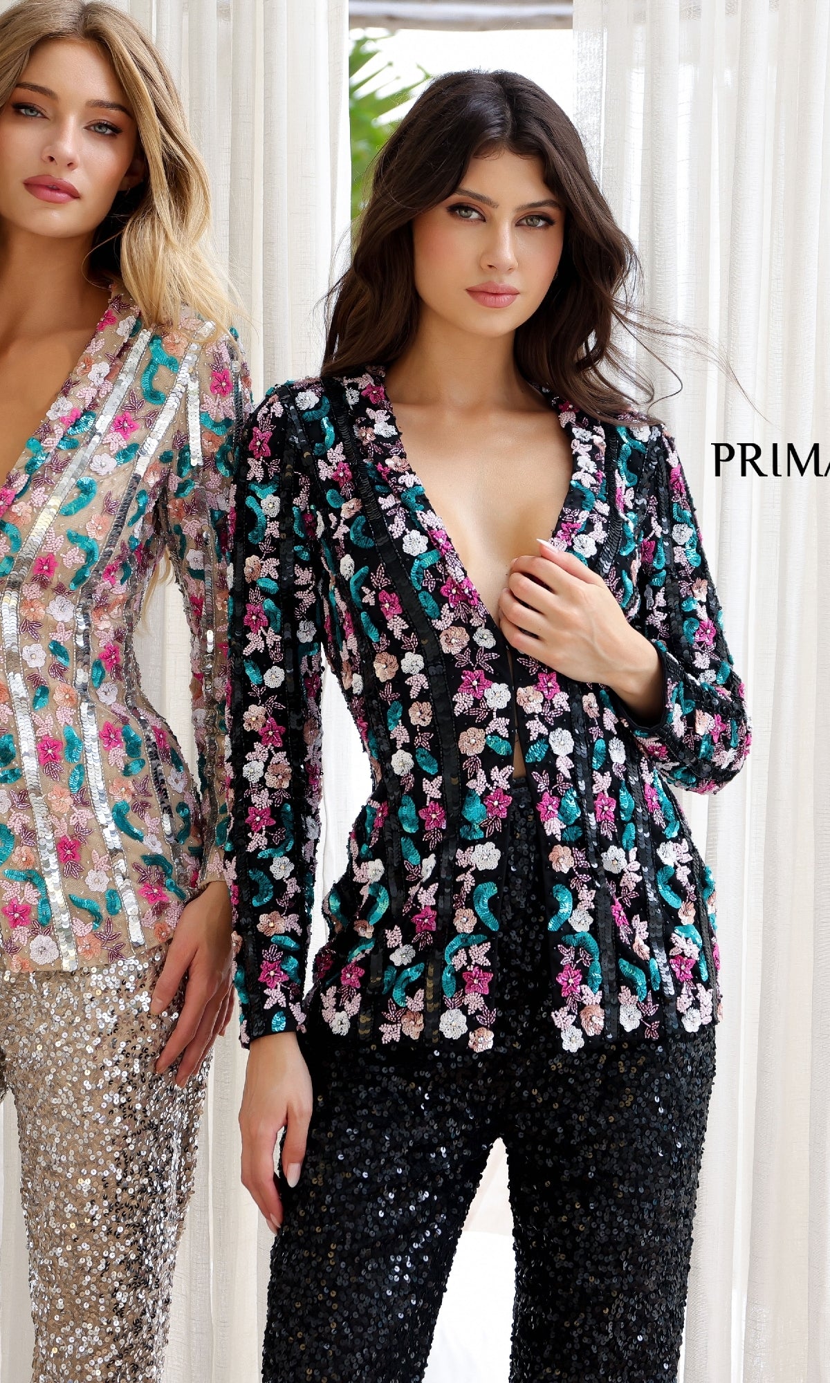 Two women stand side by side in Primavera 4388 long sequin formal jumpsuits and floral embroidered blazers—one wears black with pink and teal flowers, the other white with multicolored blooms.